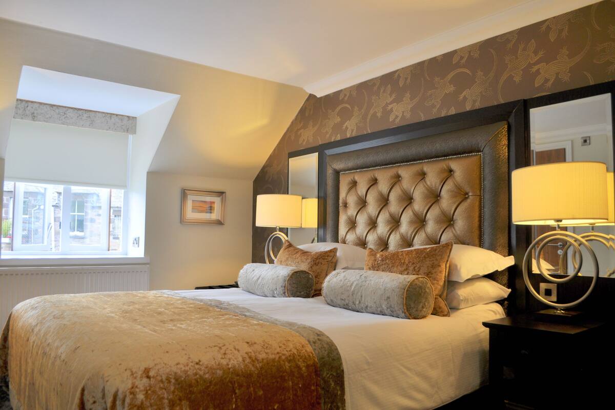 A hotel room with a large bed, two bedside lamps, a headboard with a tufted design, and a window with a view of buildings.