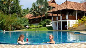 A swimming pool with a tiled edge, surrounded by lush greenery and a building with a tiled roof in the background.
