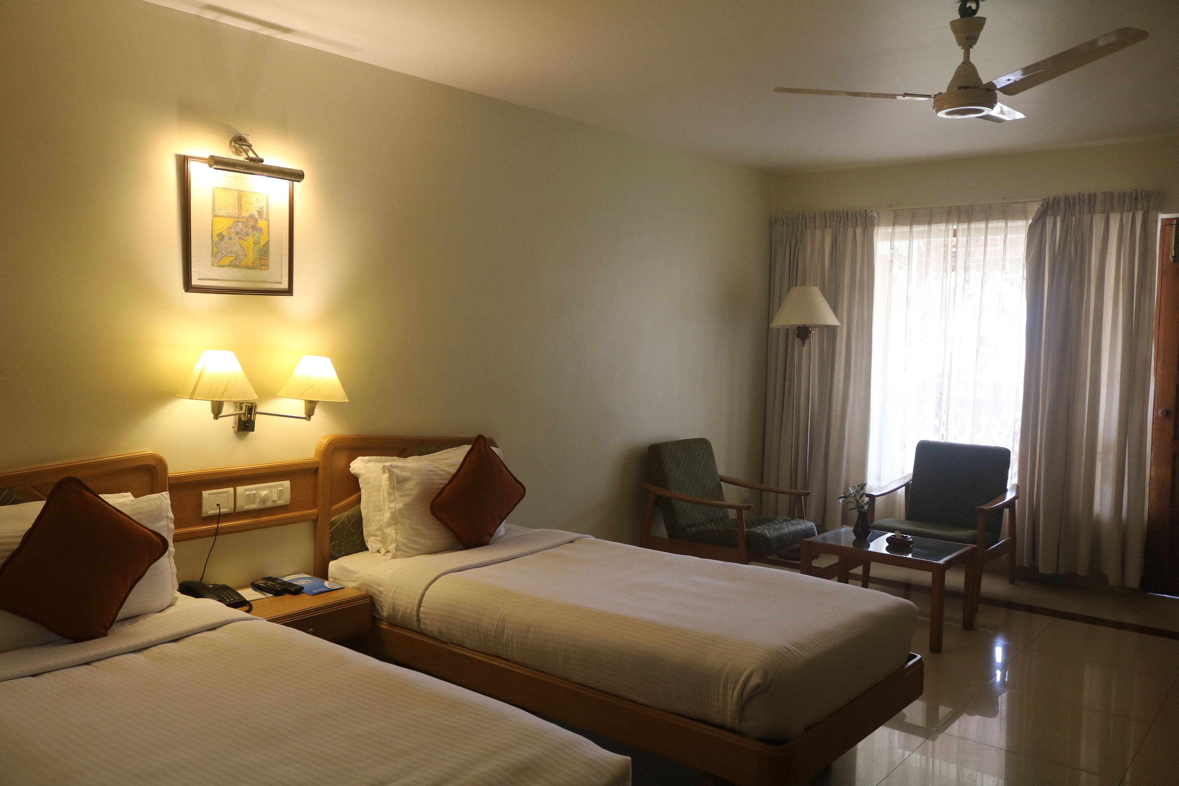 A hotel room with two beds, a ceiling fan, a desk with a chair, a lamp, and a window with curtains.