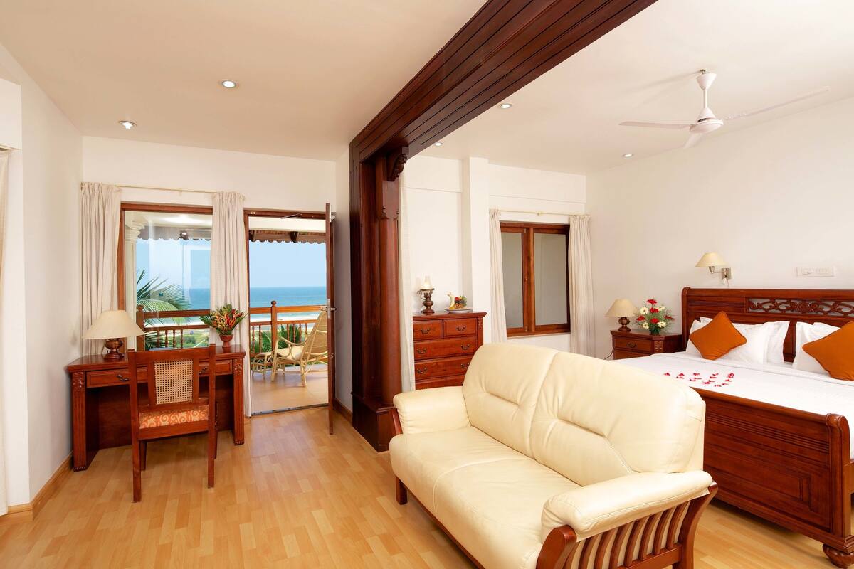 A hotel room with a wooden bed, a leather sofa, a desk with a lamp, and a view of the ocean through a glass door.