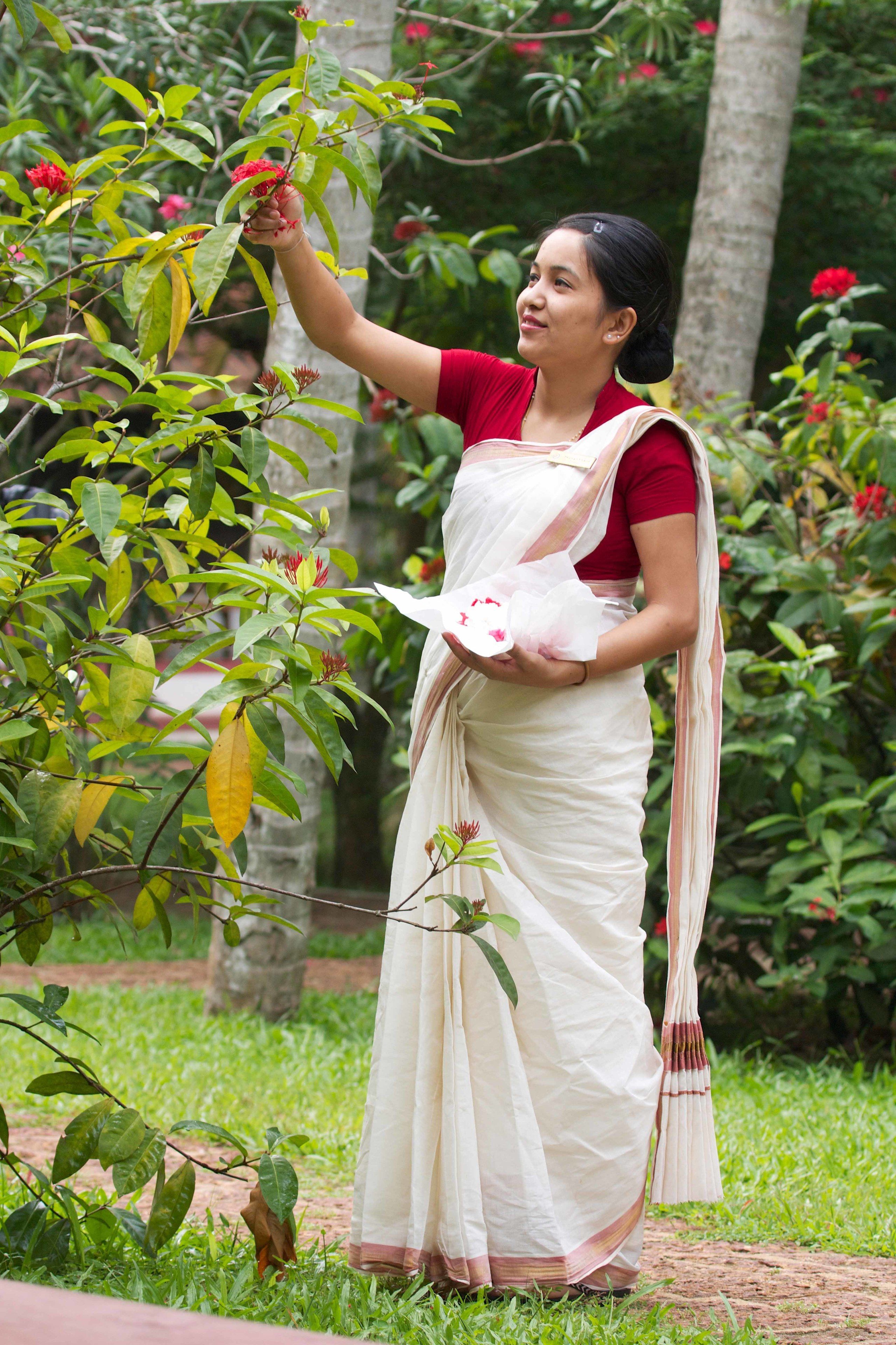 Una persona vestida con atuendo tradicional parada en un jardín con abundante follaje y plantas florecientes.