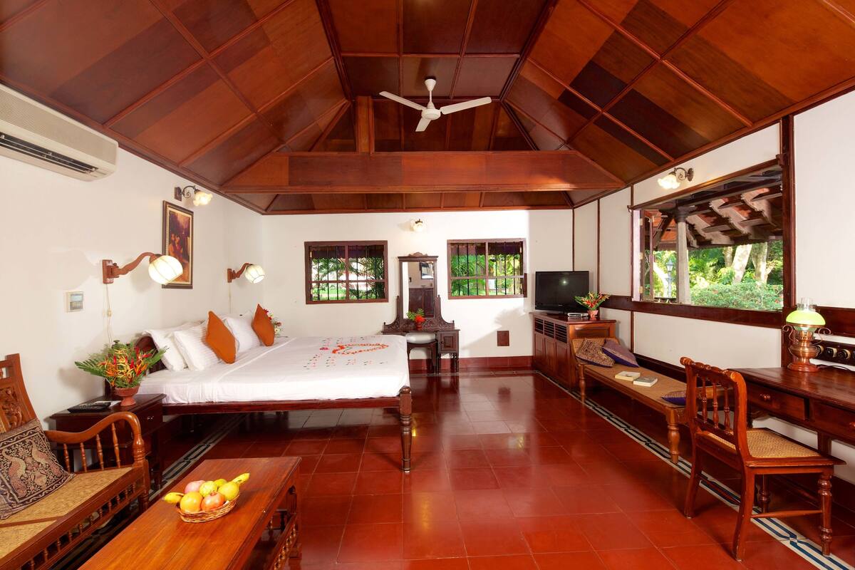 A spacious room with a bed, a desk, and a chair. There is a ceiling fan and a window with a view of greenery outside.