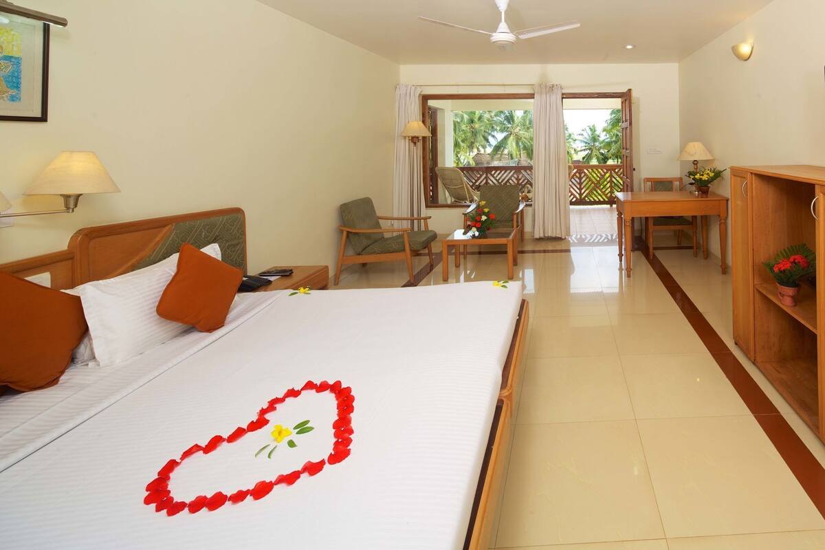 A hotel room with a bed, a desk, a chair, and a balcony with a view of palm trees.
