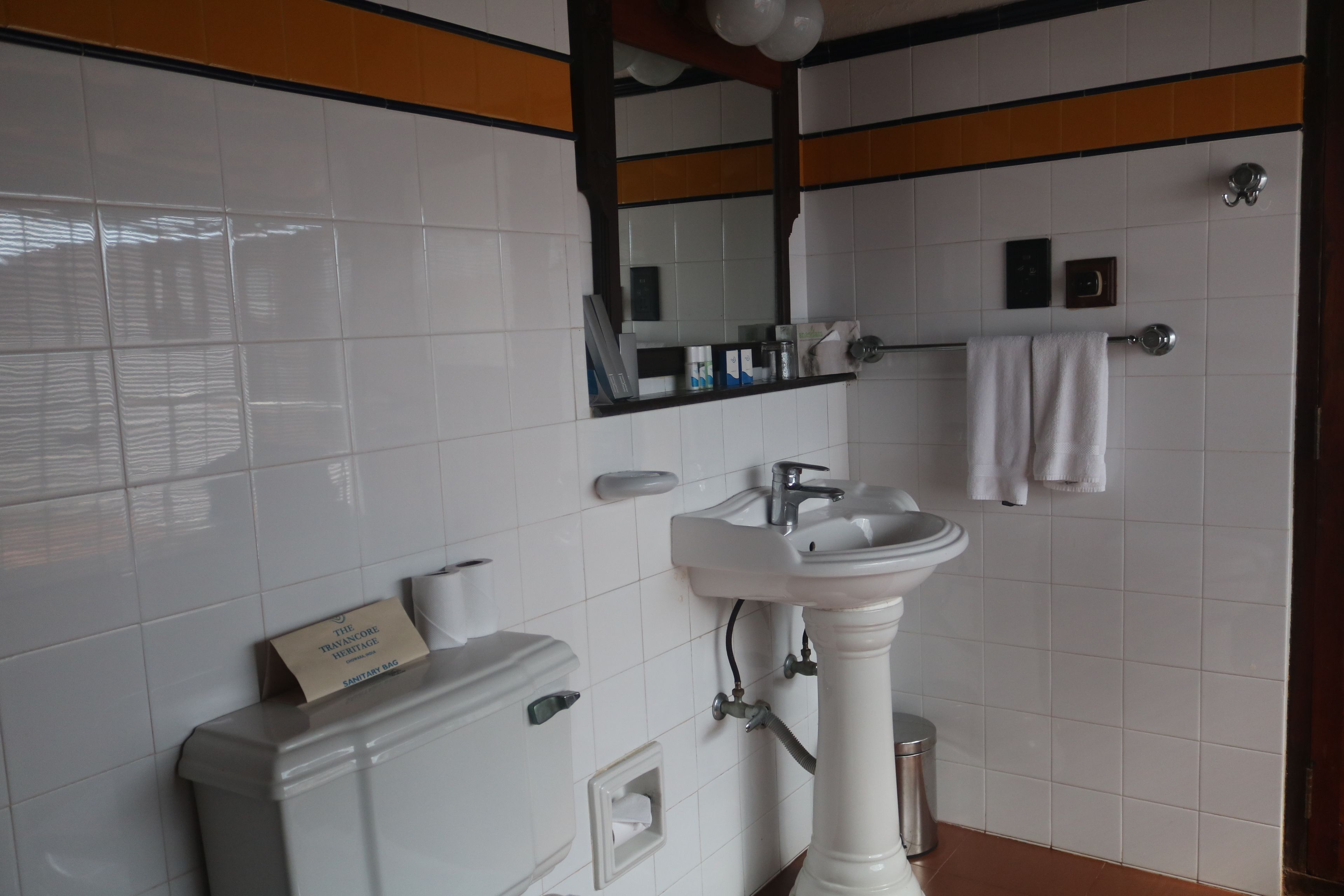 Un baño con azulejos blancos, un lavamanos pedestal, un inodoro, un espejo y un perchero para toallas.
