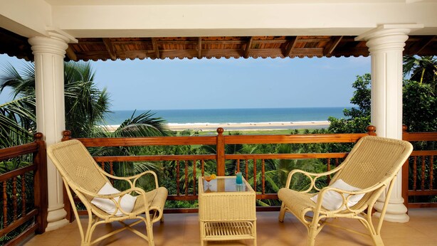 A balcony with wicker chairs and a small table, overlooking a beach and ocean.