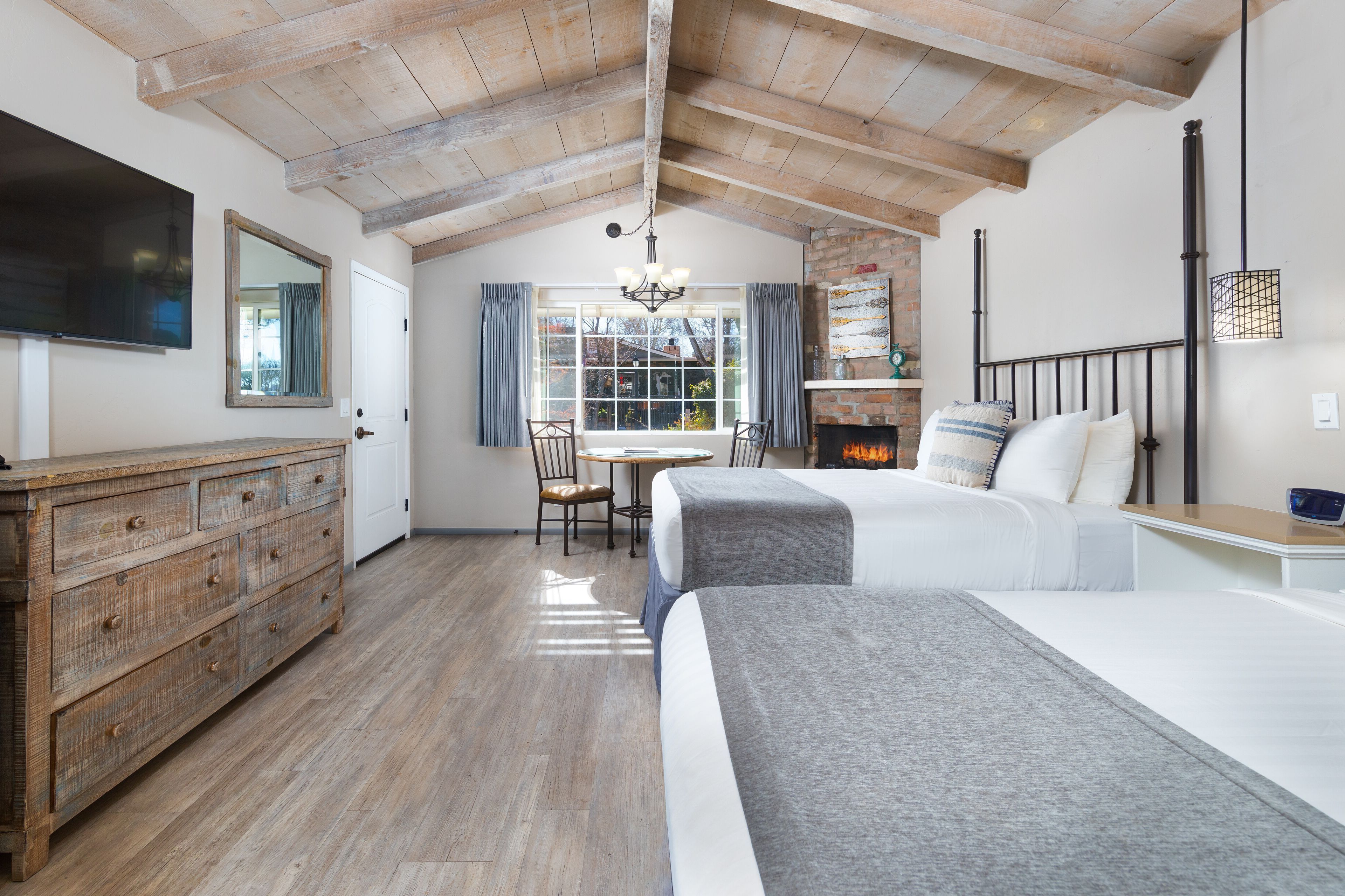 A bedroom with a wooden ceiling, a bed, a dresser, a TV, a window with curtains, and a fireplace.