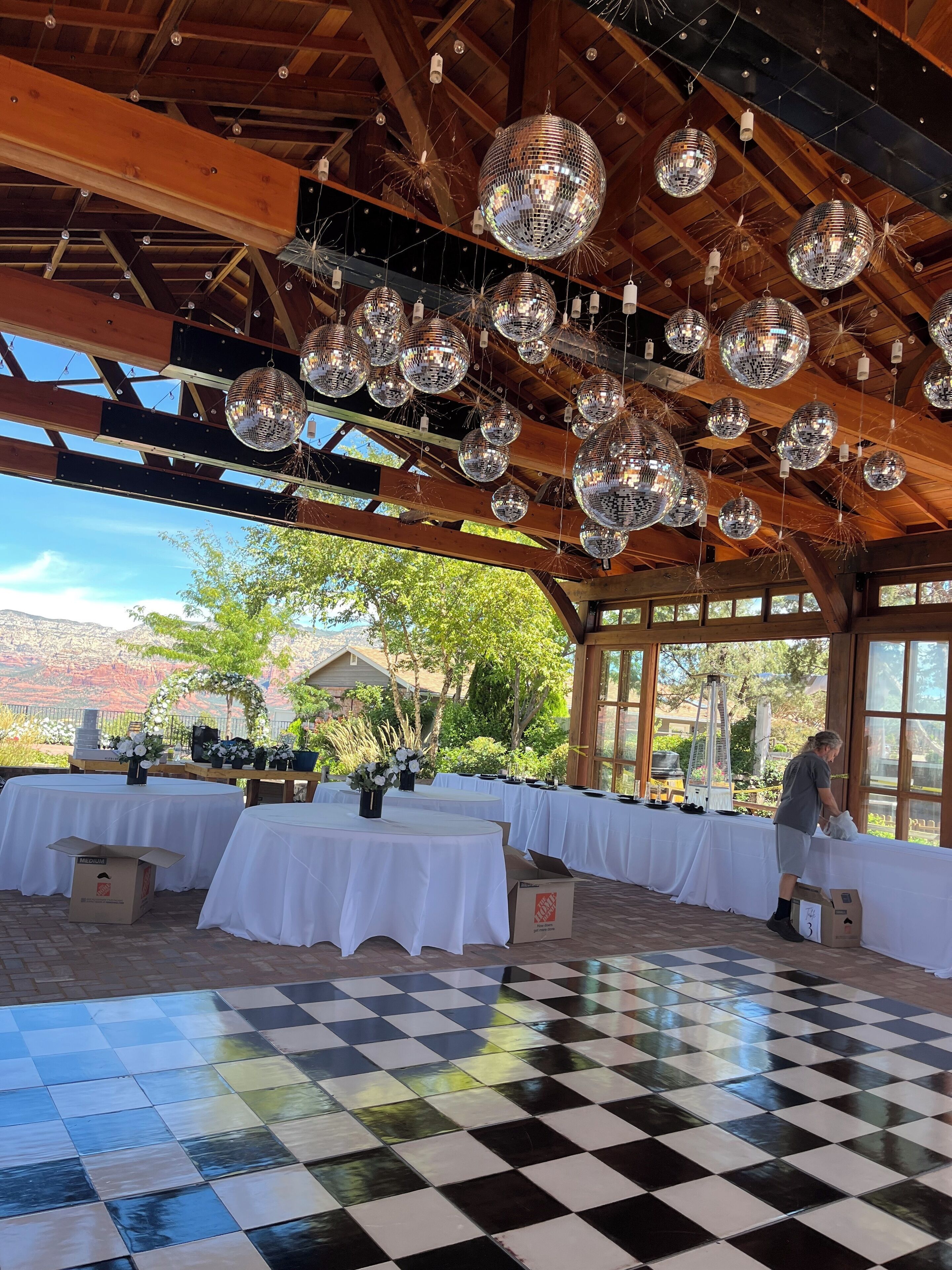 Un salón amplio con piso a cuadros, vigas de madera y bolas de discoteca colgando del techo. Se han dispuesto mesas con manteles blancos y centros de mesa florales.