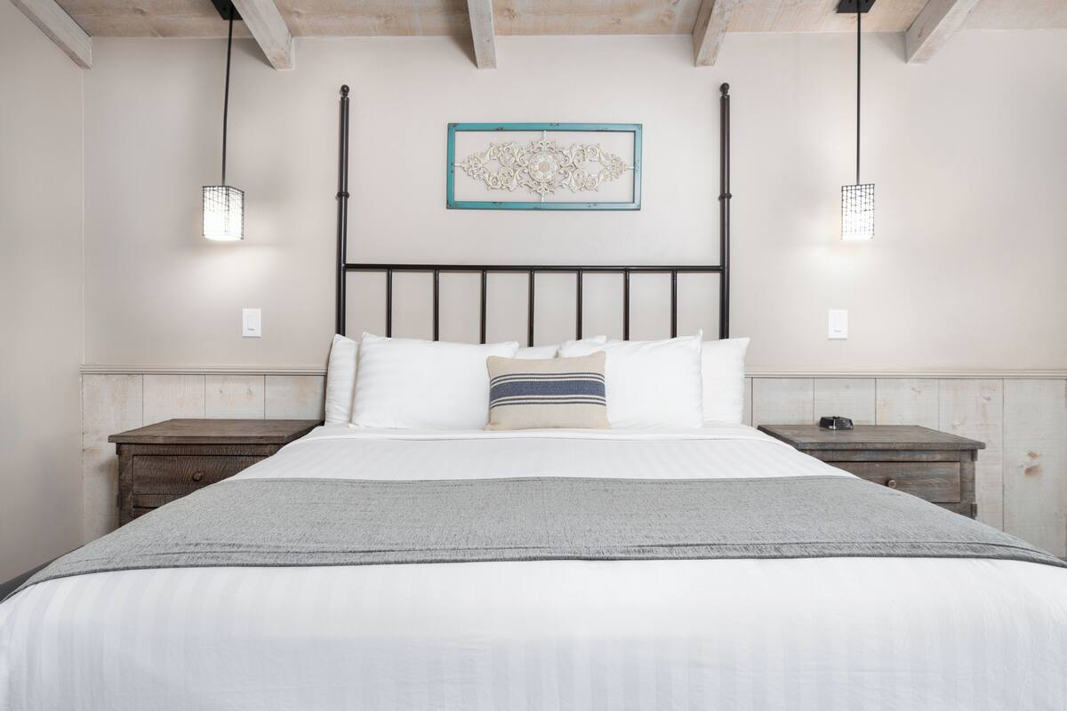 Cama bem arrumada com manta cinza, duas mesas de cabeceira e um objeto decorativo na parede.