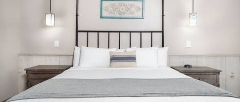 Cama bem arrumada com lençóis brancos e manta cinza, acompanhada por duas mesas de cabeceira de madeira com uma pequena luminária em cada.