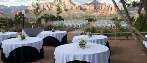 Round tables with white tablecloths and black bases, set up outdoors with a mountainous backdrop.