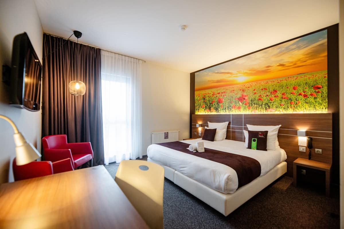 Une chambre d’hôtel avec un grand lit, un bureau, une chaise rouge, un tableau représentant un champ et une lampe.