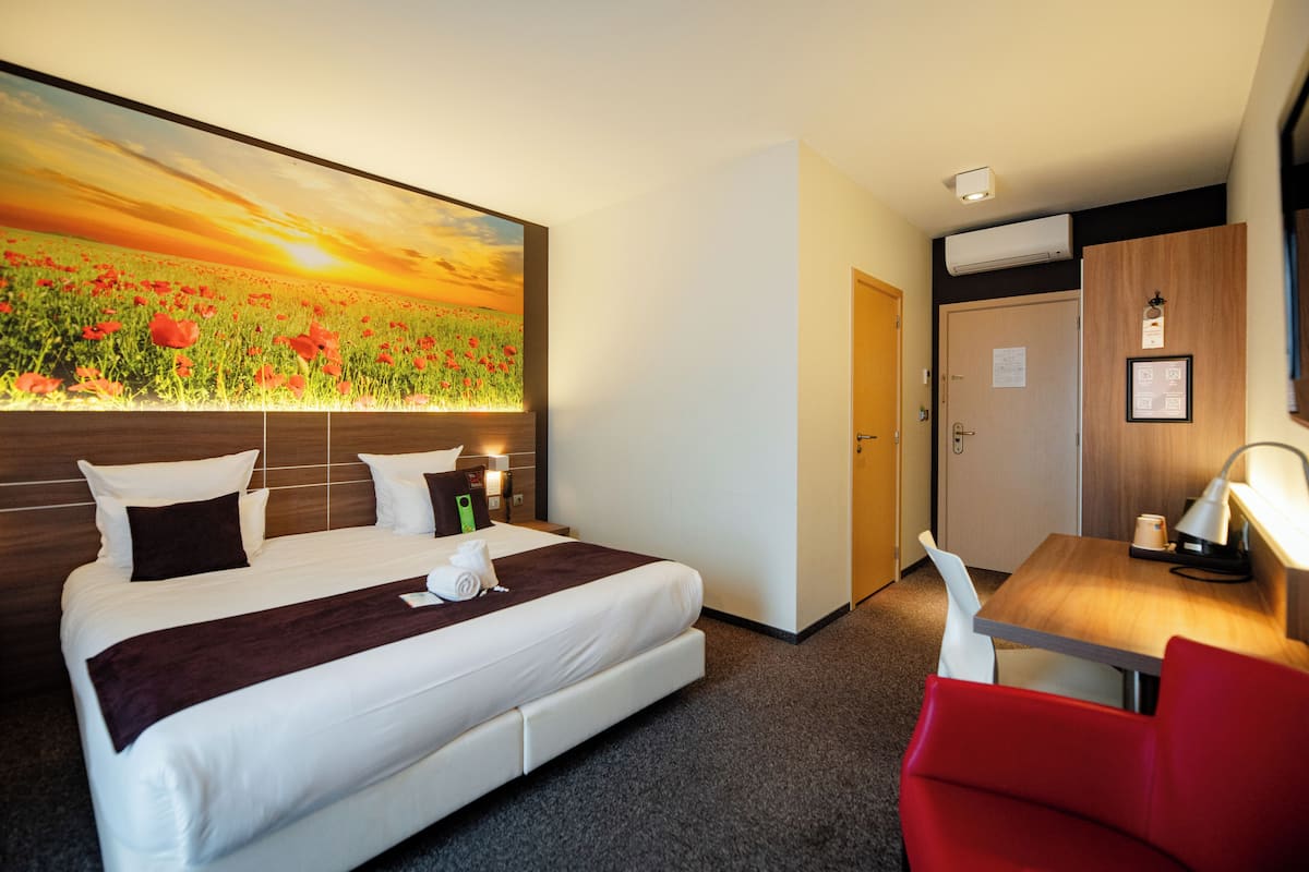 Une chambre d’hôtel avec un grand lit, un bureau avec une chaise, un tableau représentant un champ et une porte.