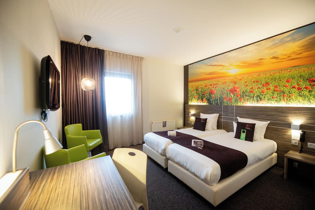 Une chambre d’hôtel avec un grand lit, un bureau, une chaise et un tableau représentant un champ parsemé de fleurs rouges.