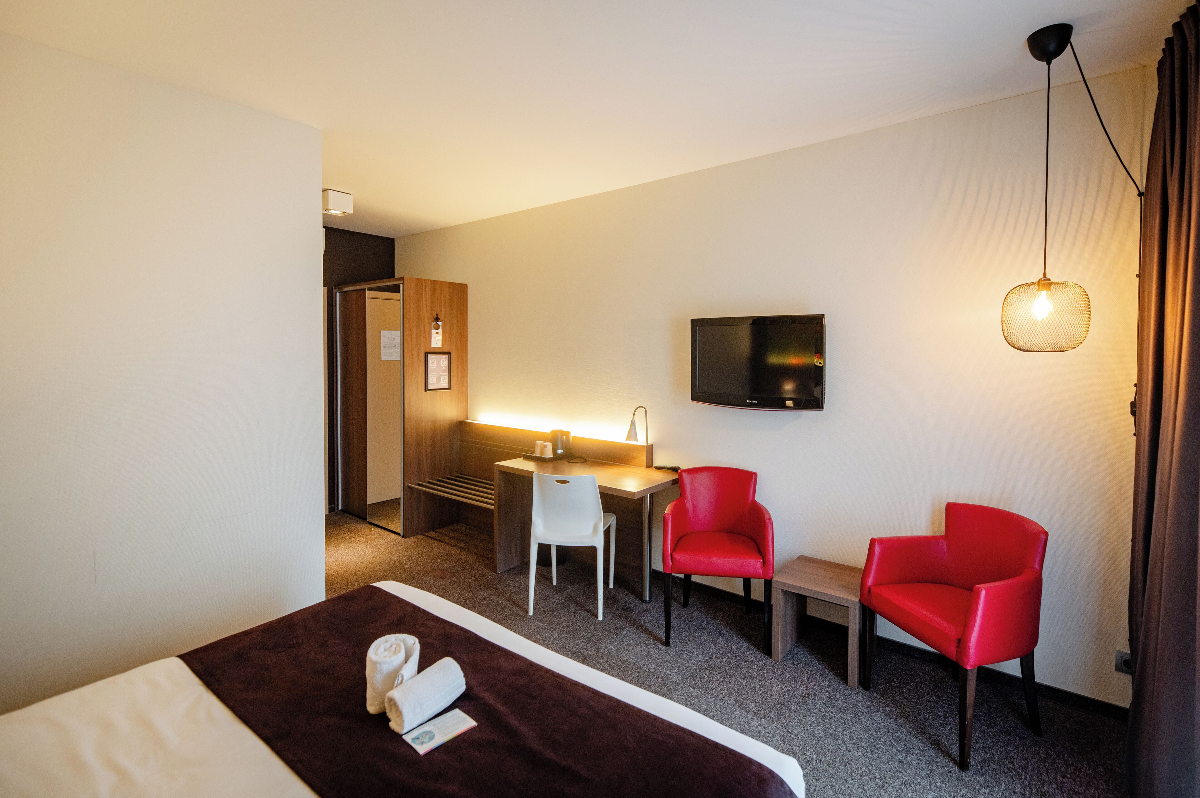 Habitación de hotel con cama, escritorio, dos sillas rojas, televisor y lámpara colgante.