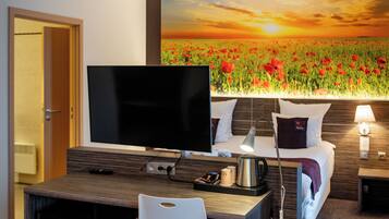 Een hotelkamer met een bed, een televisie, een bureau met een stoel, een koffiezetapparaat en een schilderij van een zonsondergang boven een bloemenveld.