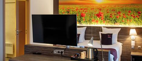 Habitación de hotel con cama, televisor, escritorio con silla, cafetera y un cuadro de una puesta de sol sobre un campo de amapolas.