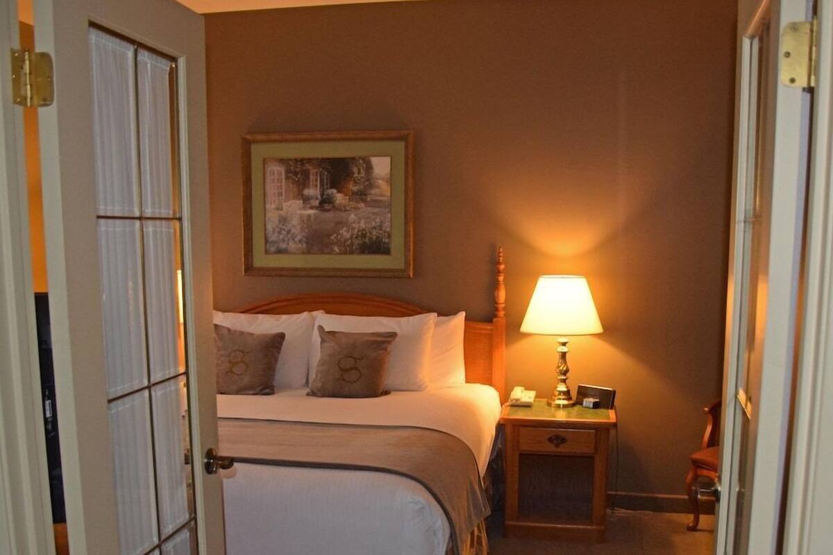 A hotel room with a bed, a nightstand with a lamp, a framed picture on the wall, and a door leading to another room.