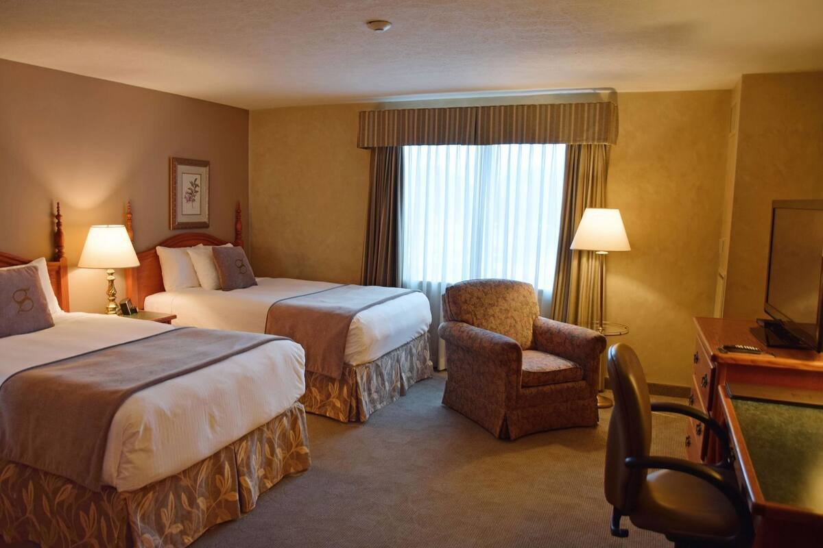 A hotel room with two beds, a chair, a desk with a television, and a window with curtains.