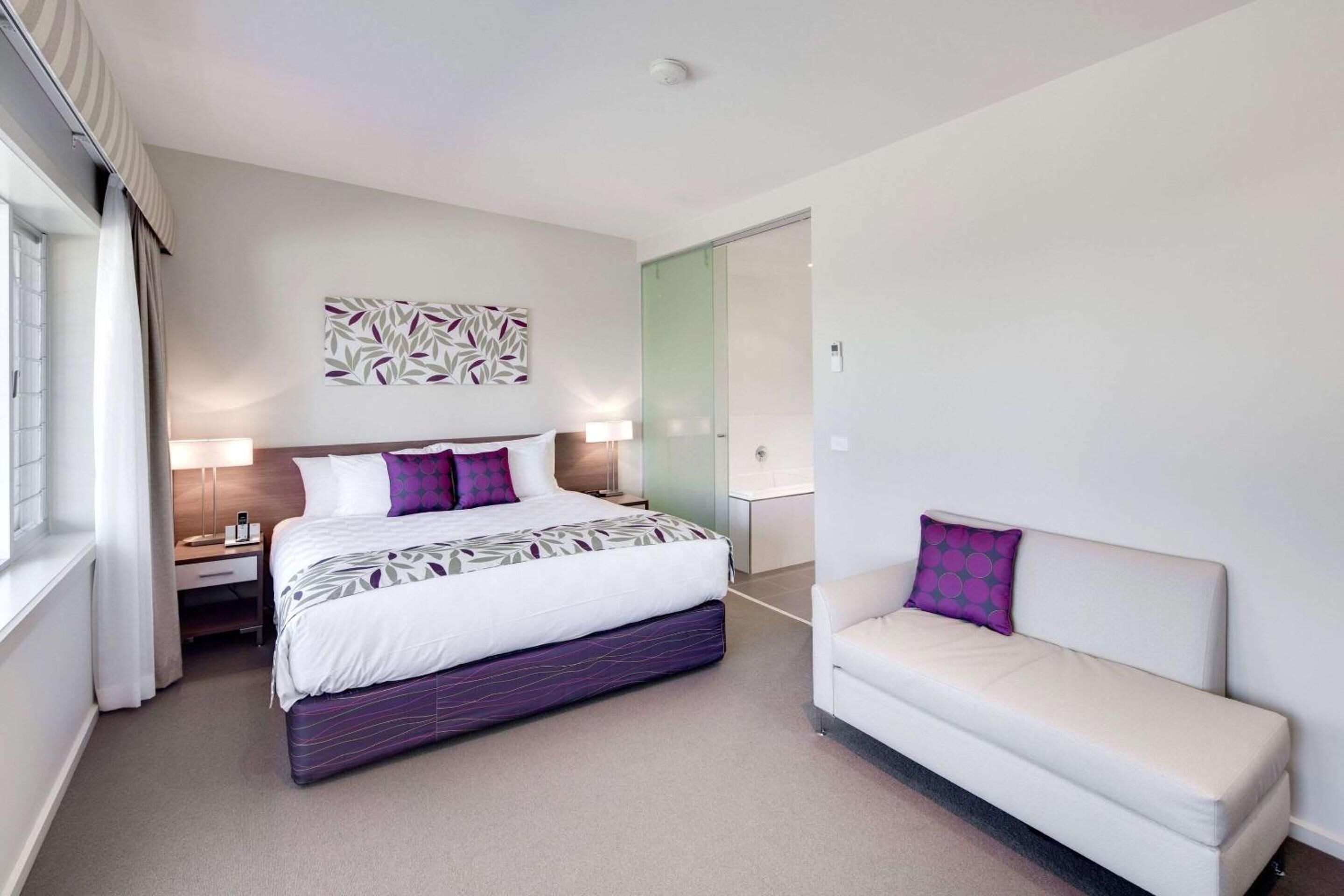 A modern hotel room with a bed, bedside table, lamp, and a sofa.