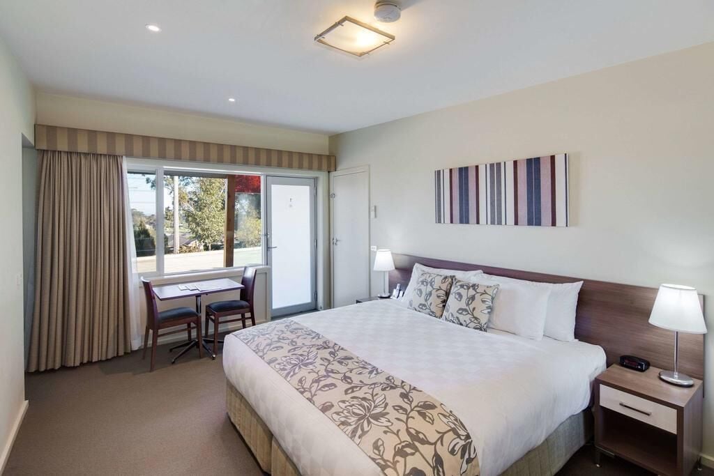 A hotel room with a large bed, a desk with two chairs, a bedside table with a lamp, and a view of the outdoors through a sliding door.