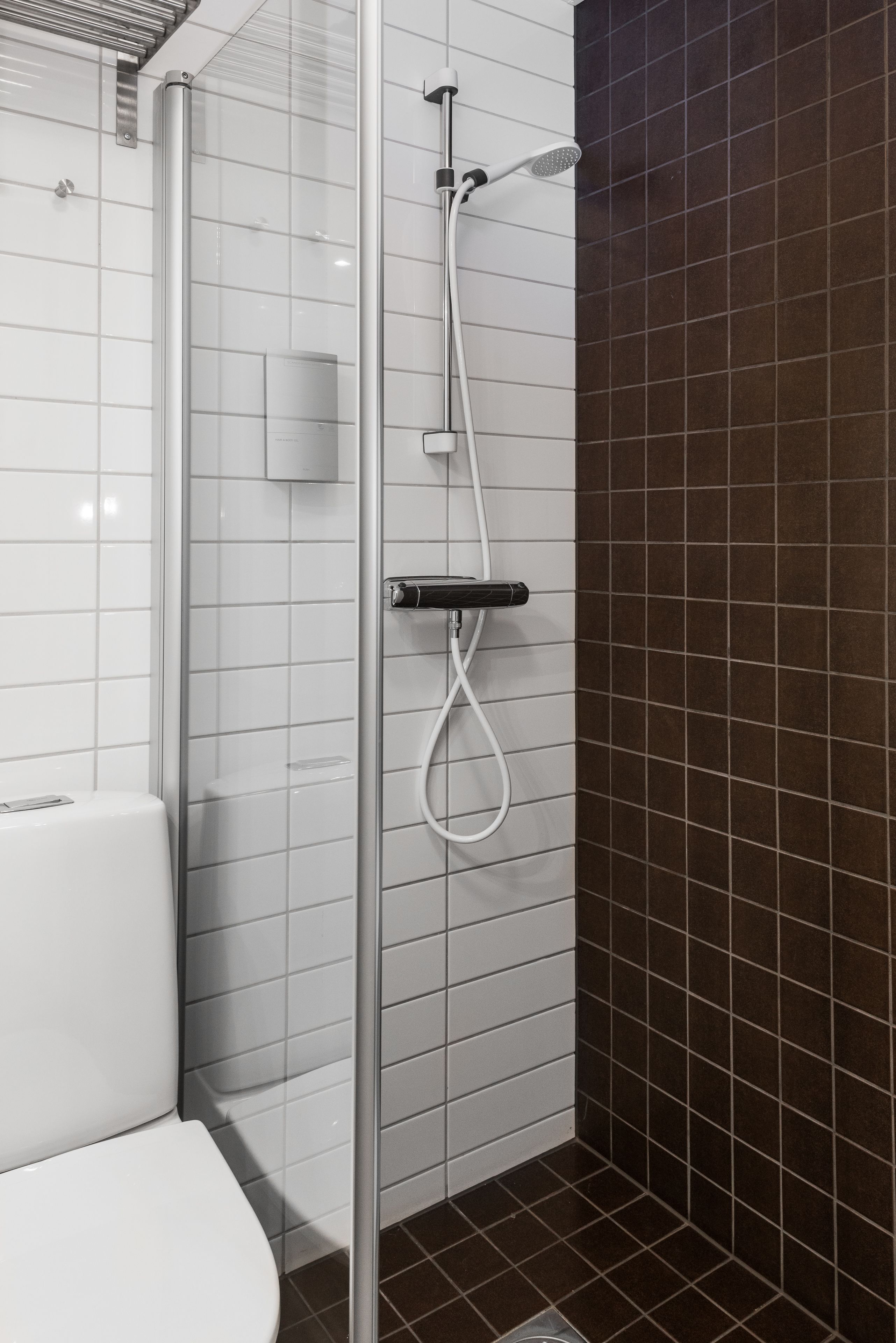 Une salle de bain moderne avec une douche à parois de verre, des toilettes blanches et des murs recouverts de carreaux de couleur brun foncé.