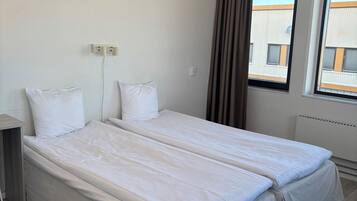 Habitación de hotel con dos camas individuales, una mesita de noche, toma de corriente, ventana con cortinas y vistas a un edificio.