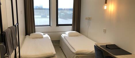 Habitación de hotel con dos camas individuales, un escritorio con silla y vista a un estacionamiento a través de dos ventanas.