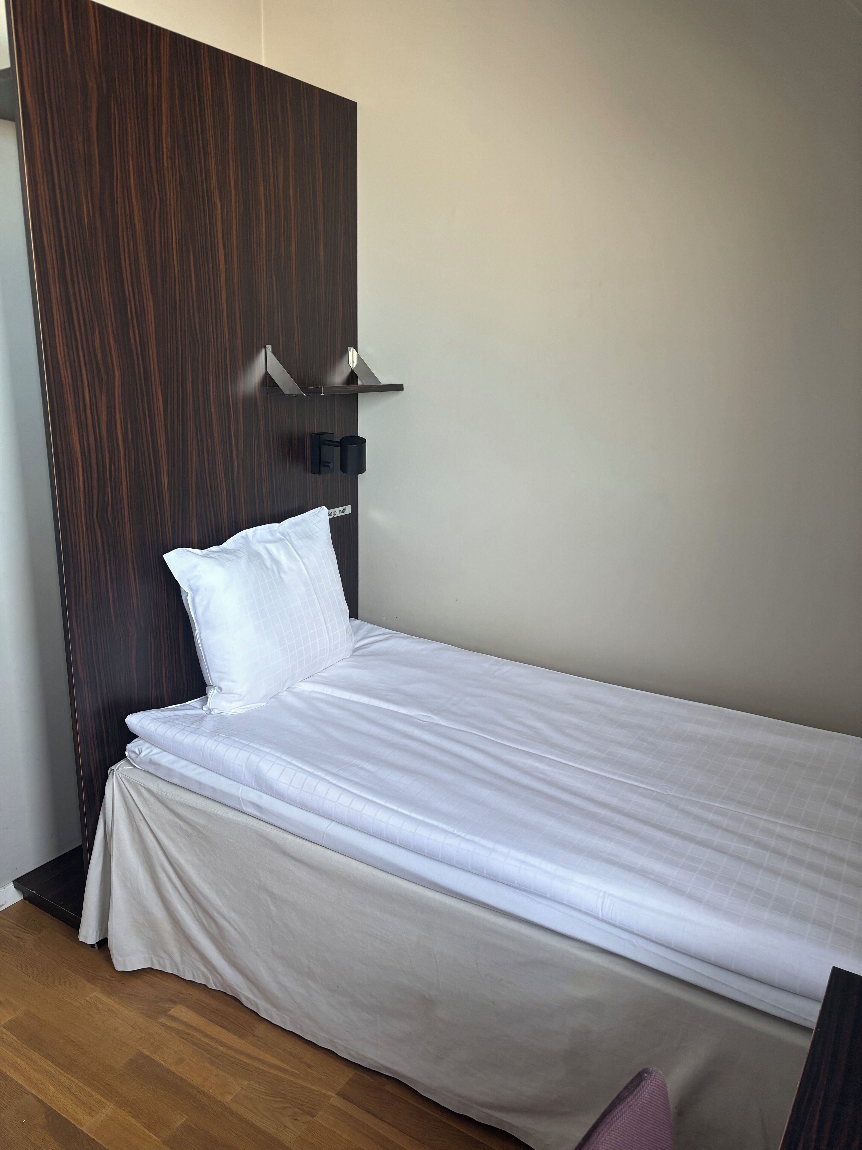 Una cama individual con cabecera de madera, una mesita de noche y un aplique de pared.