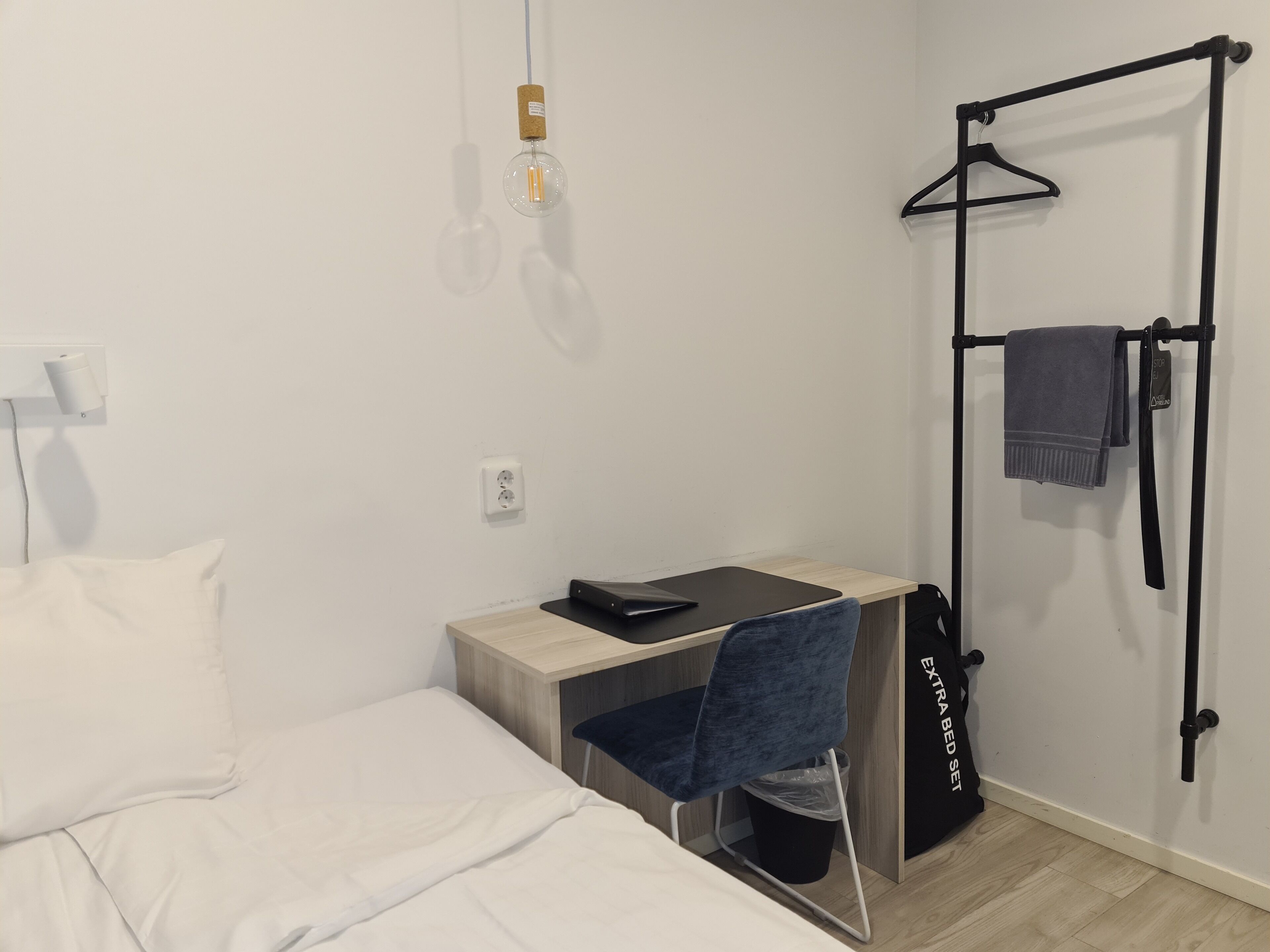 Ett kompakt hotellrum med en säng, ett skrivbord med en stol, en hängande lampa, en svart galge med en handduk och en svart väska märkt "EXTRASÄNG".