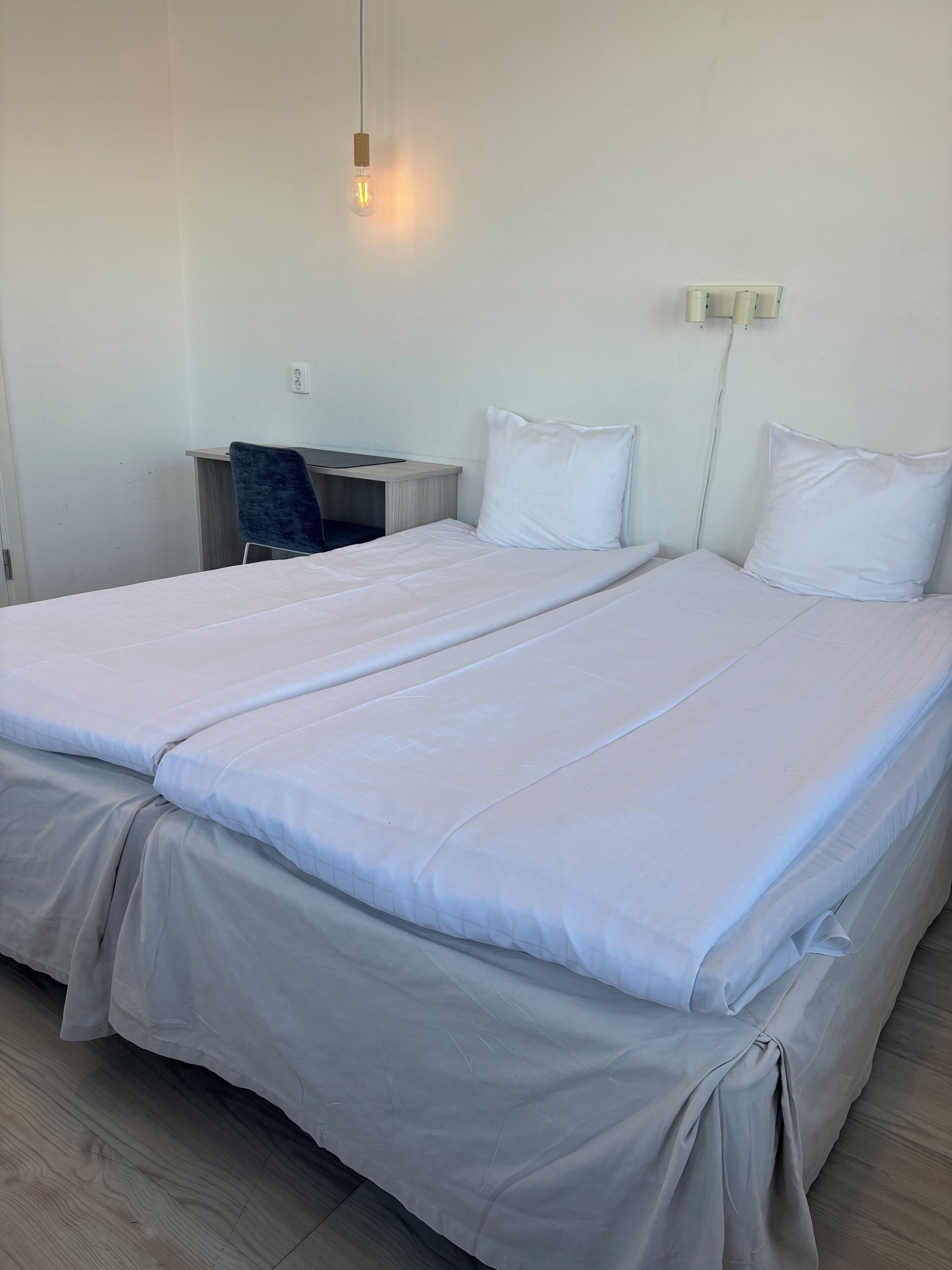A double bed with white linens, a desk with a chair, and a wall-mounted light fixture.