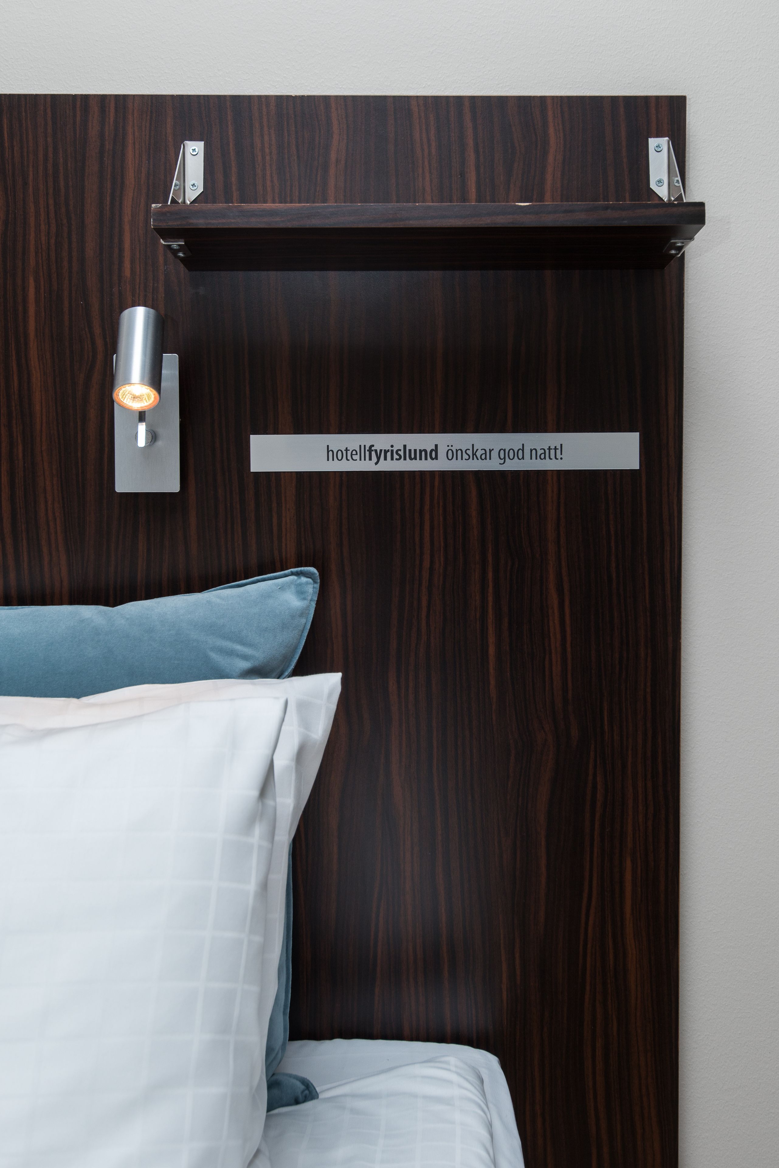 A hotel room with a dark wood headboard, a wall-mounted light fixture, and a sign that reads "hotelfyrlisland önskar god natt!"
