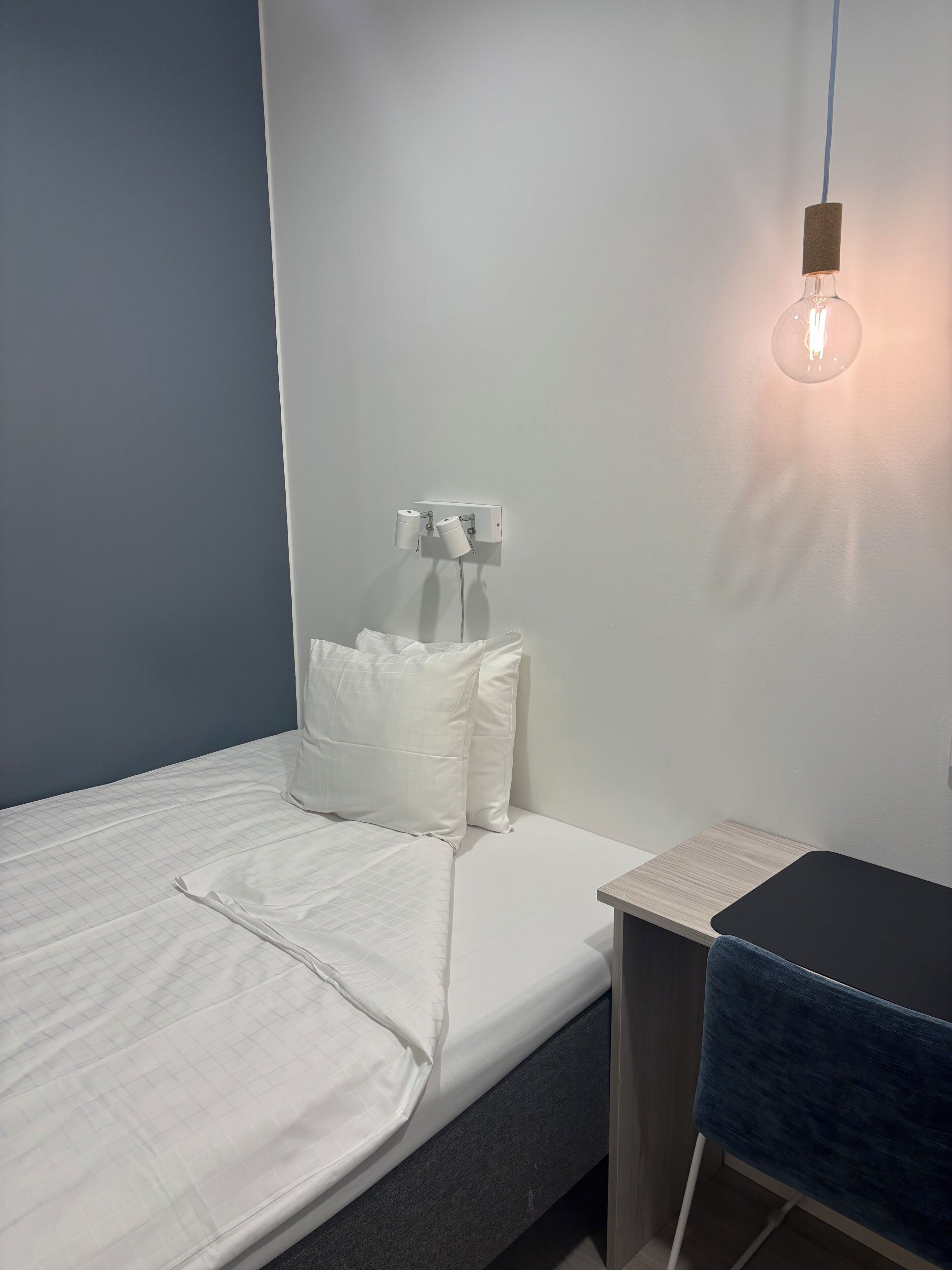Una cama bien hecha con ropa blanca, una mesita de noche pequeña y una silla azul.