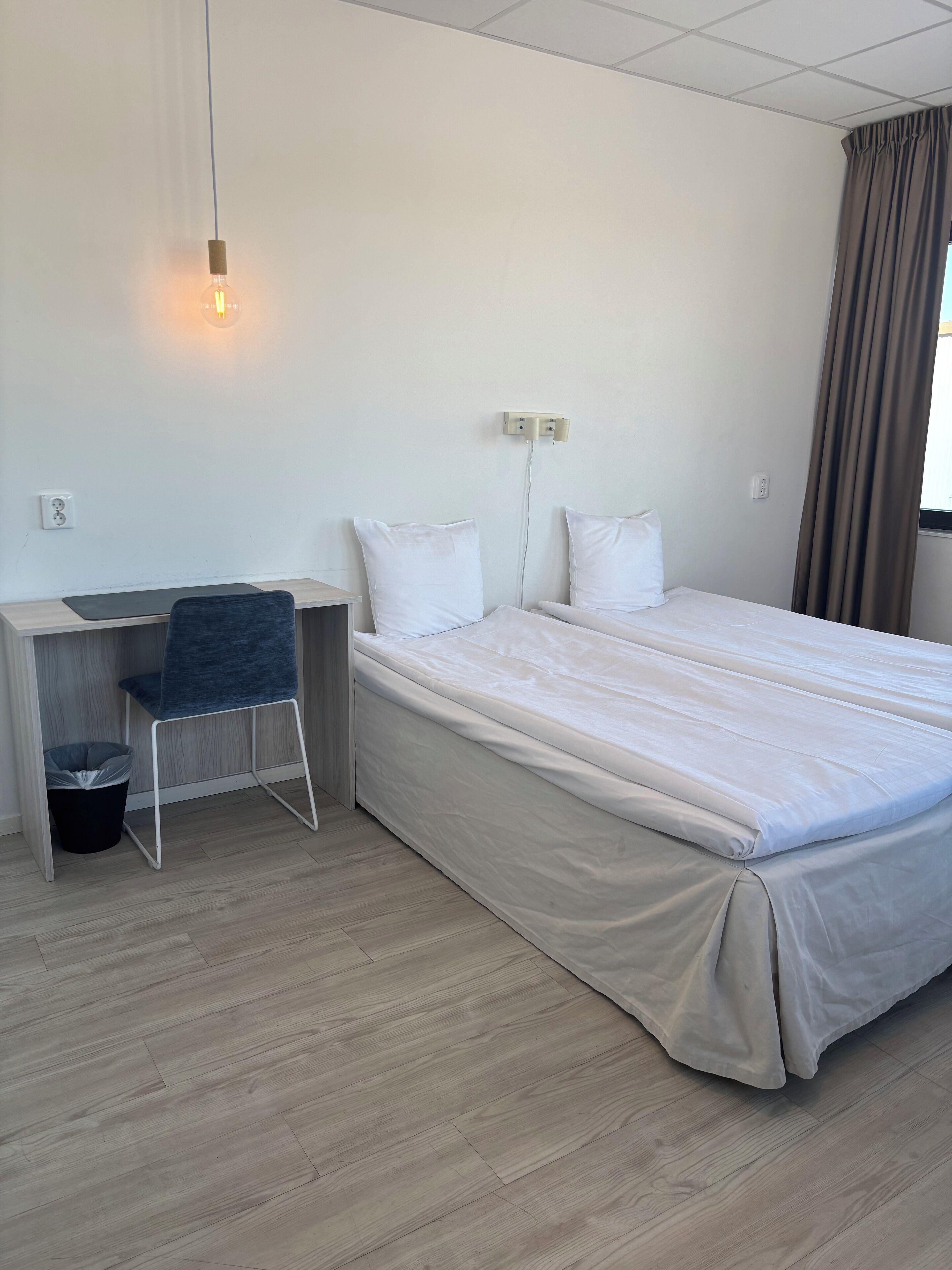 A hotel room with a bed, a desk, a chair, and a trash bin.
