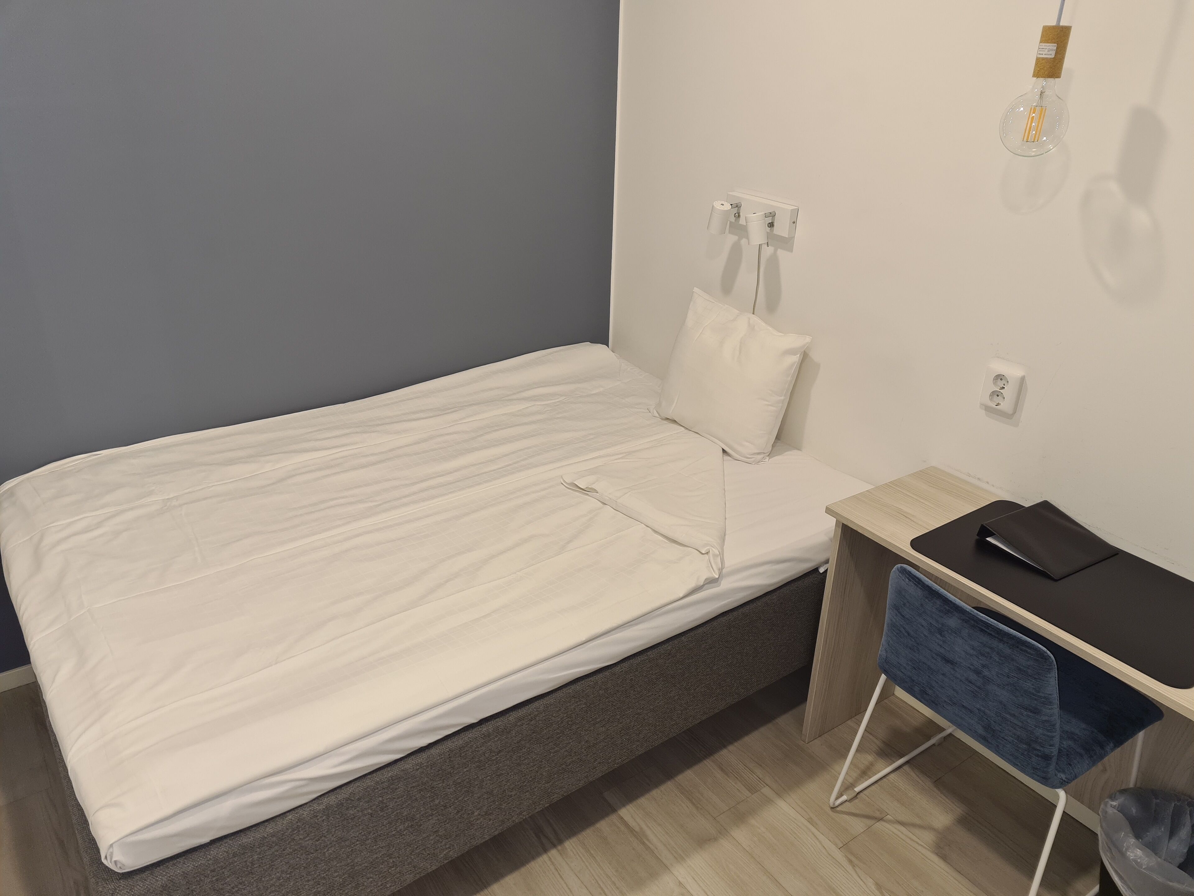 A single bed with a white mattress and pillow, a bedside table with a black surface and a blue chair, and a wall socket.