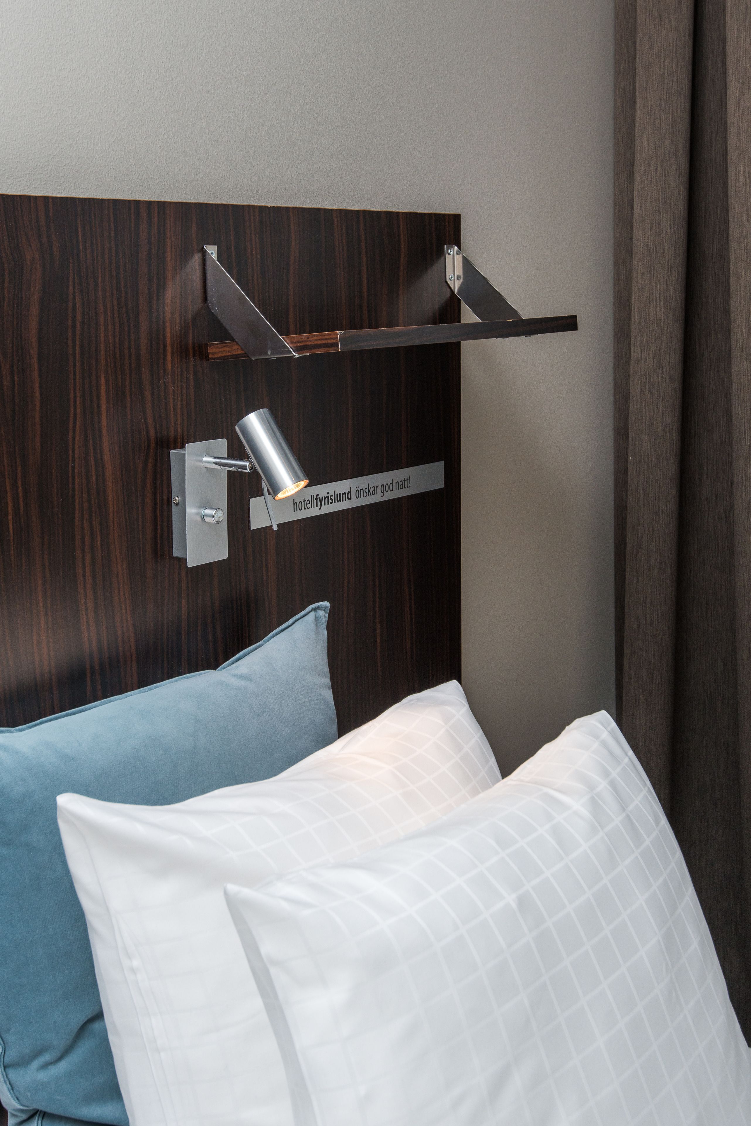 Une chambre d’hôtel avec une tête de lit en bois, une lampe de lecture fixée au mur et deux oreillers.