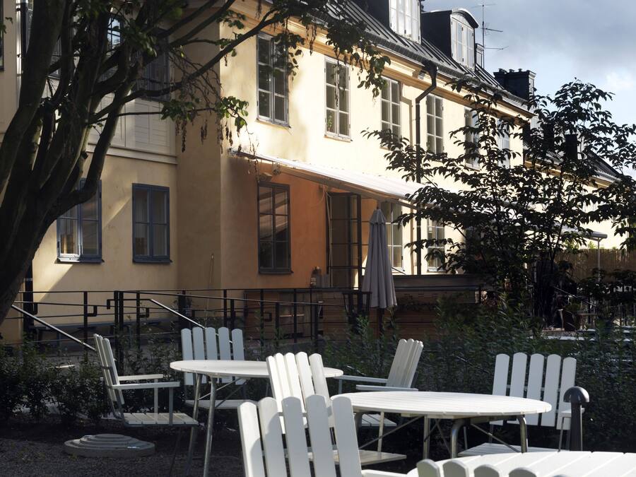 An outdoor seating area with white chairs and tables, surrounded by a building with large windows and a balcony.