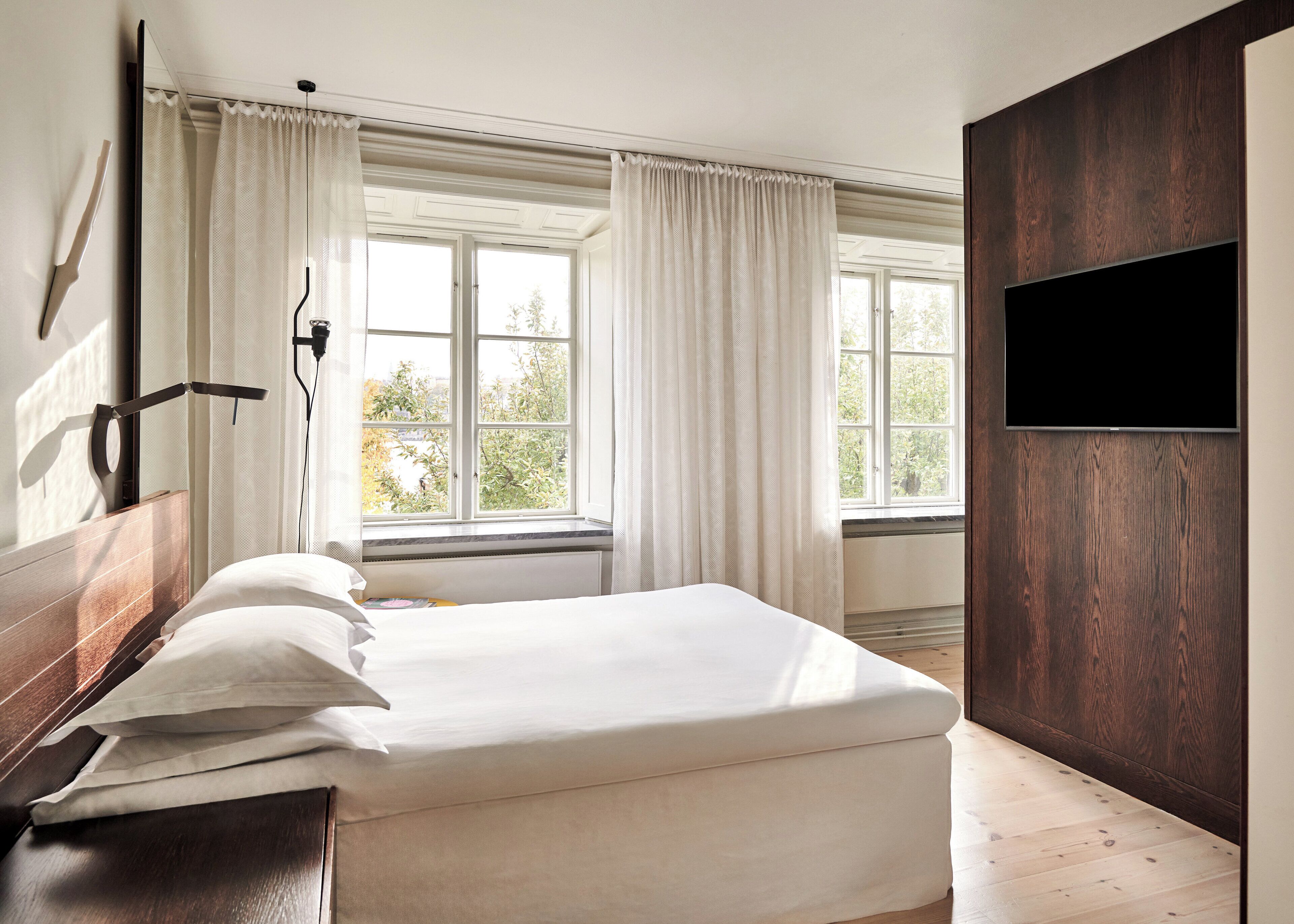 A bedroom with a bed, a window with curtains, a wall-mounted TV, and a wooden floor.