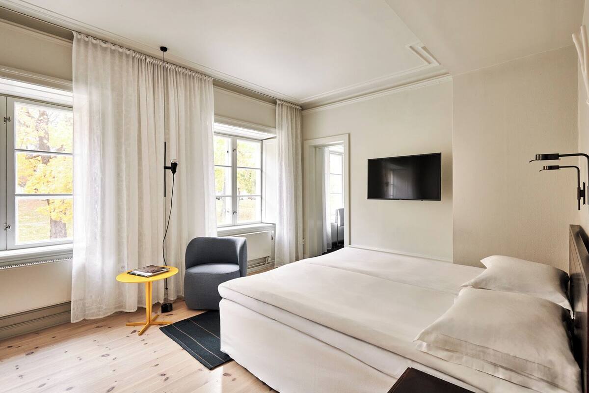 A bedroom with a large bed, a chair, a small table, and a TV on the wall.