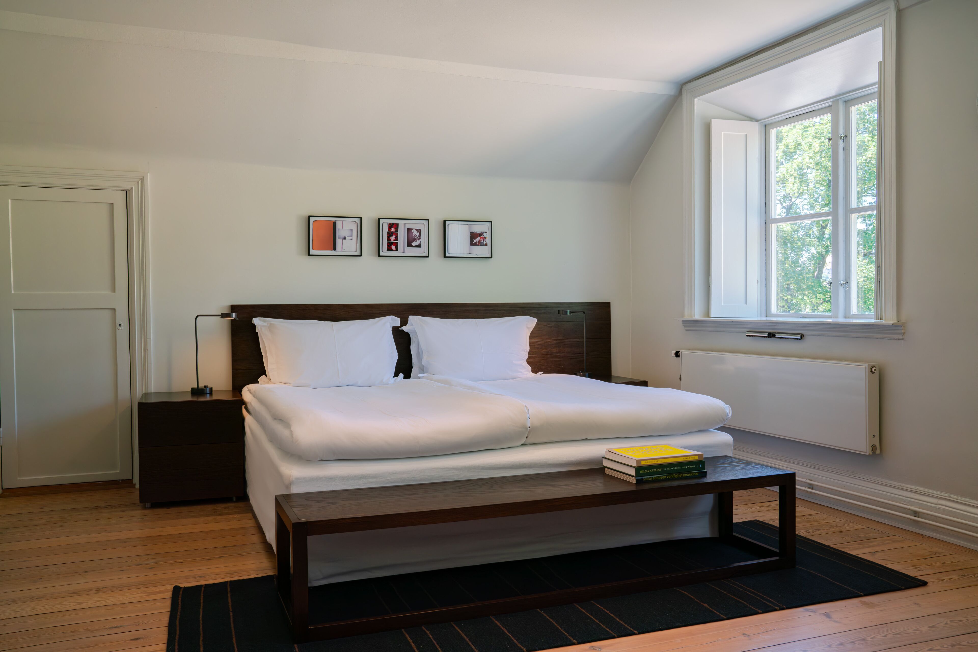 A bedroom with a bed, a nightstand, a bedside table, a window, and framed pictures on the wall.