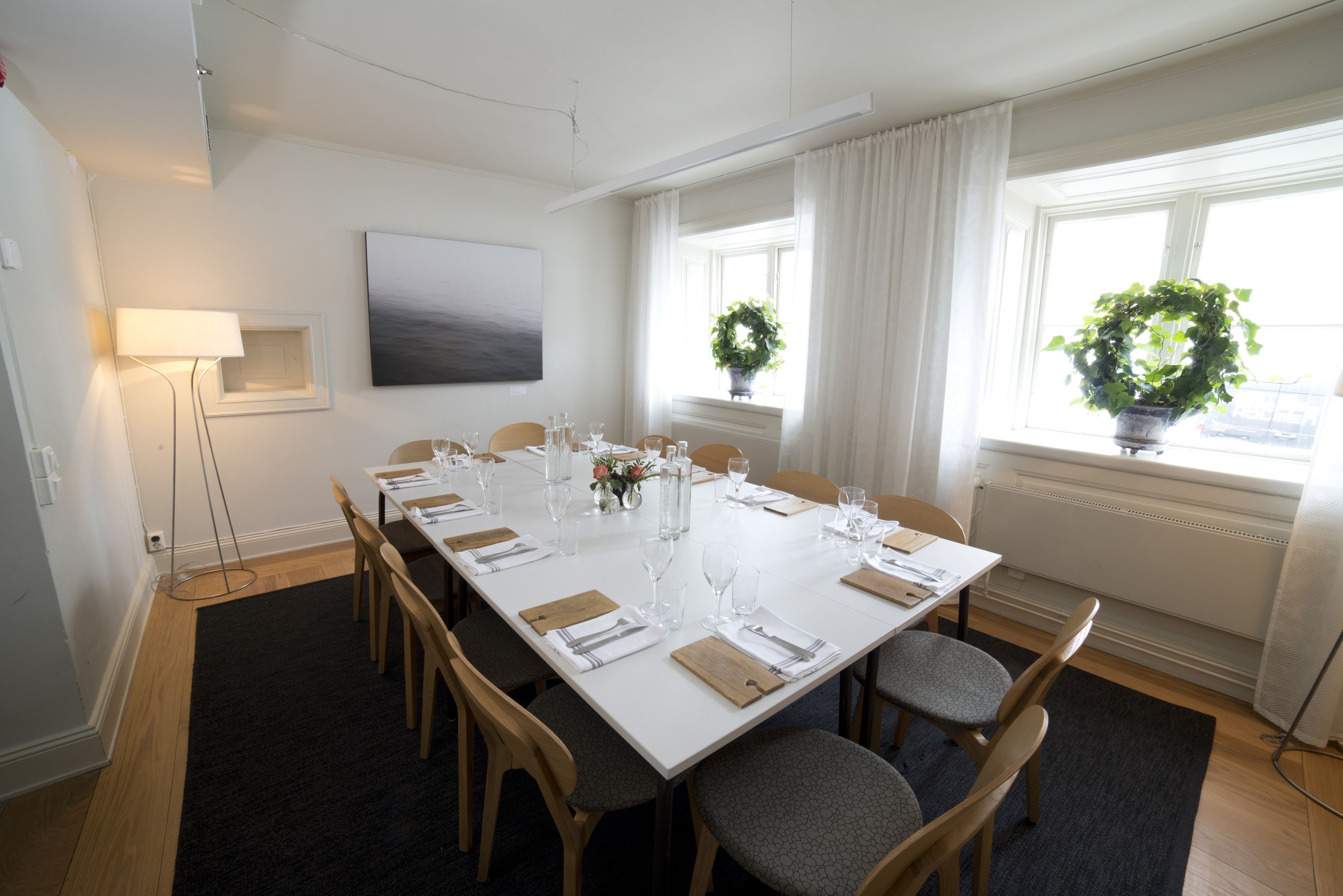 A dining room with a long table set for six, surrounded by chairs, with a floor lamp and a painting on the wall.