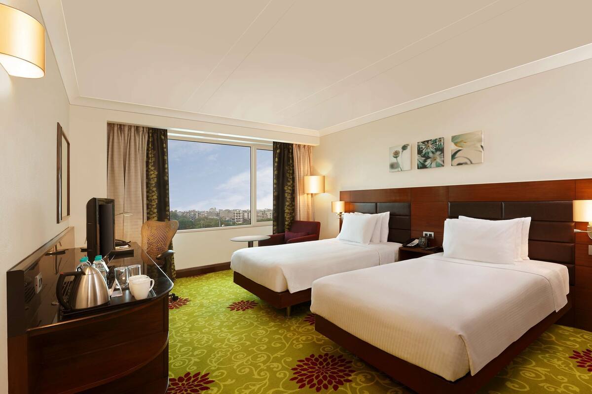 A hotel room with two beds, a desk with a kettle, a TV, and a window with curtains.