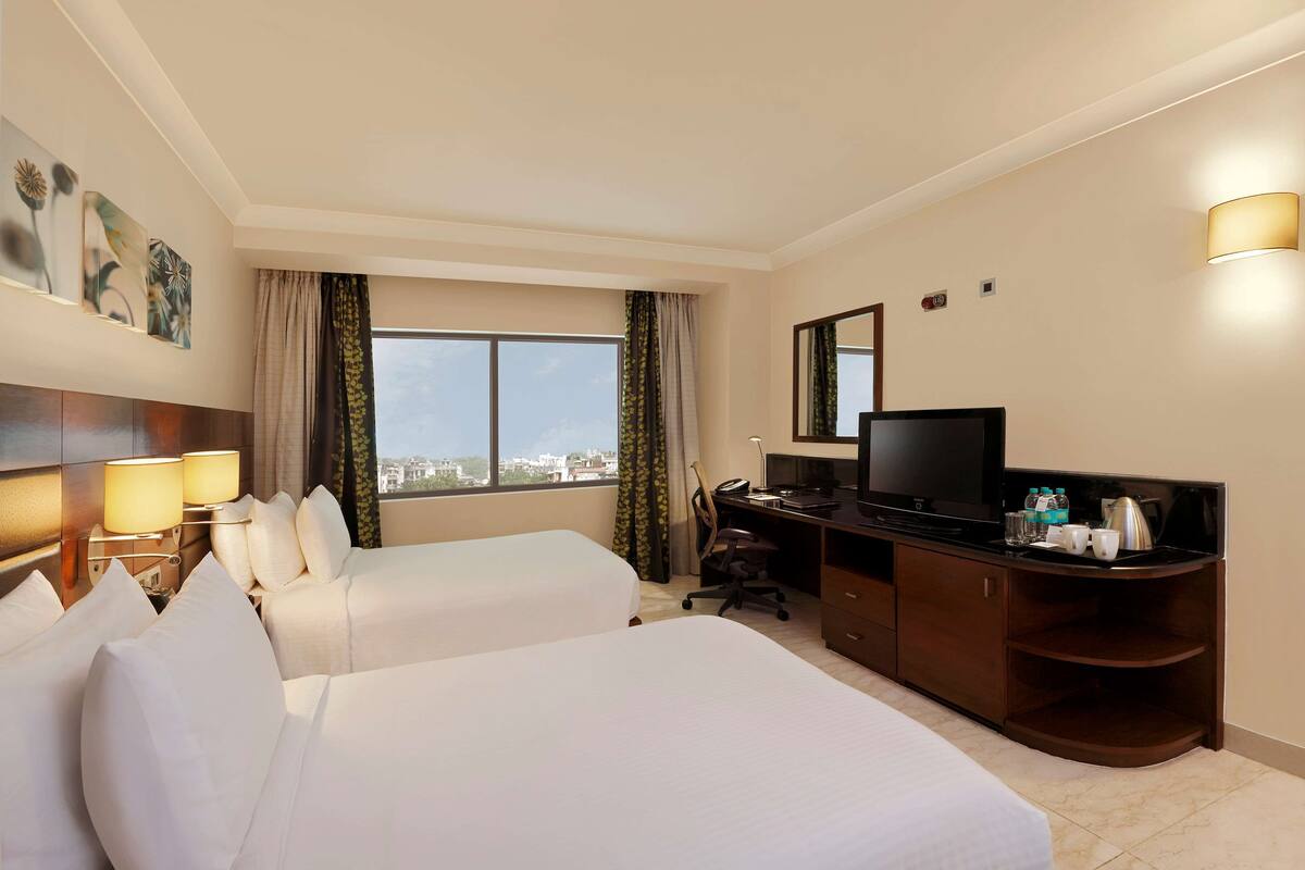A hotel room with two beds, a desk with a TV, a lamp, and a view of the city.