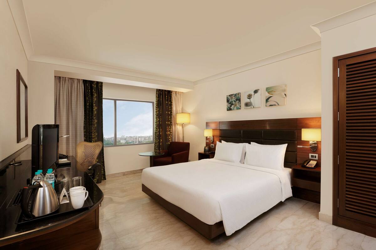 A hotel room with a large bed, a desk with a flat-screen TV, a chair, a nightstand with lamps, and a view of the city through the window.