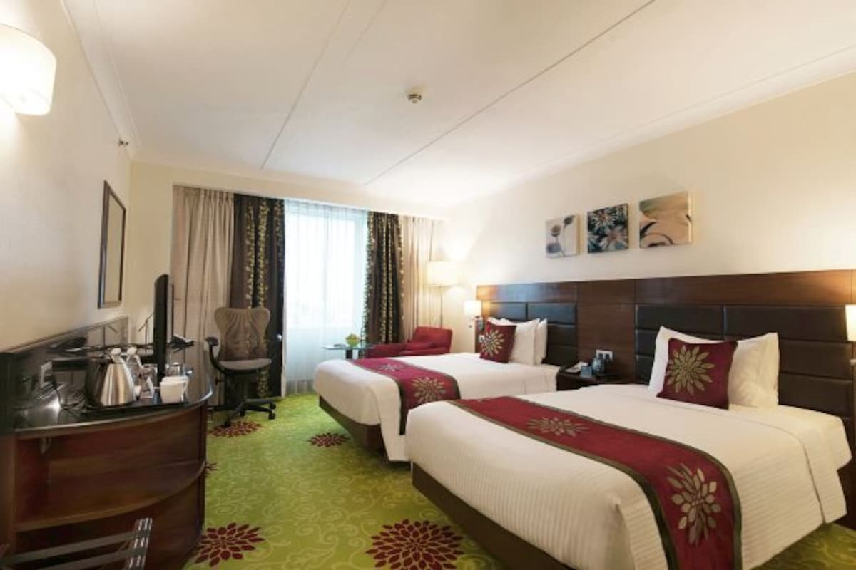 Habitación de hotel con dos camas, un escritorio con silla, una lámpara y una ventana con cortinas.