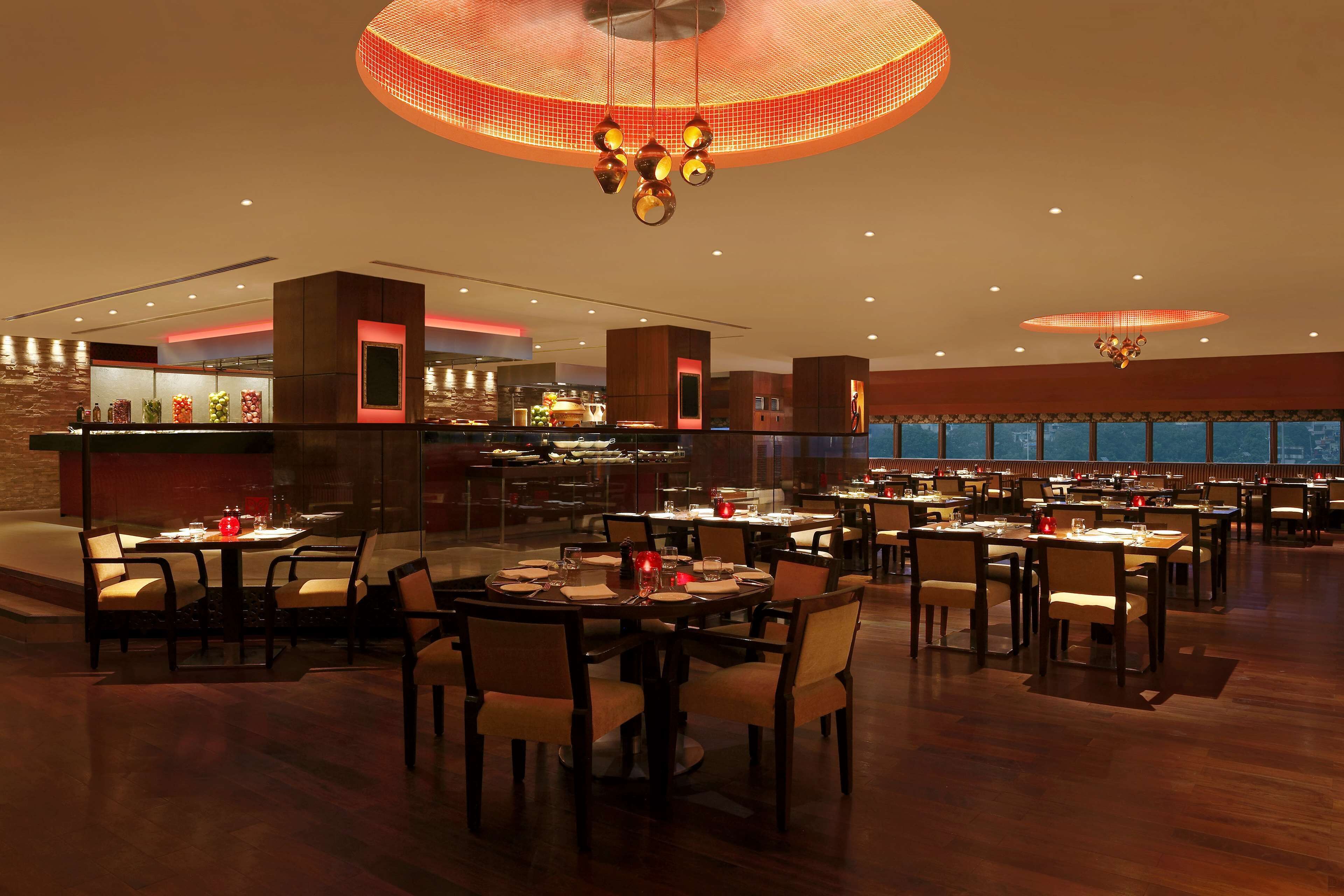 A spacious hotel dining area with multiple tables and chairs, a modern chandelier, and a view of the cityscape through large windows.