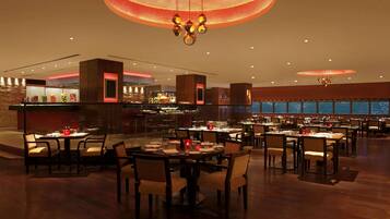 A spacious hotel dining area with multiple tables and chairs, a modern chandelier, and a view of the cityscape through large windows.