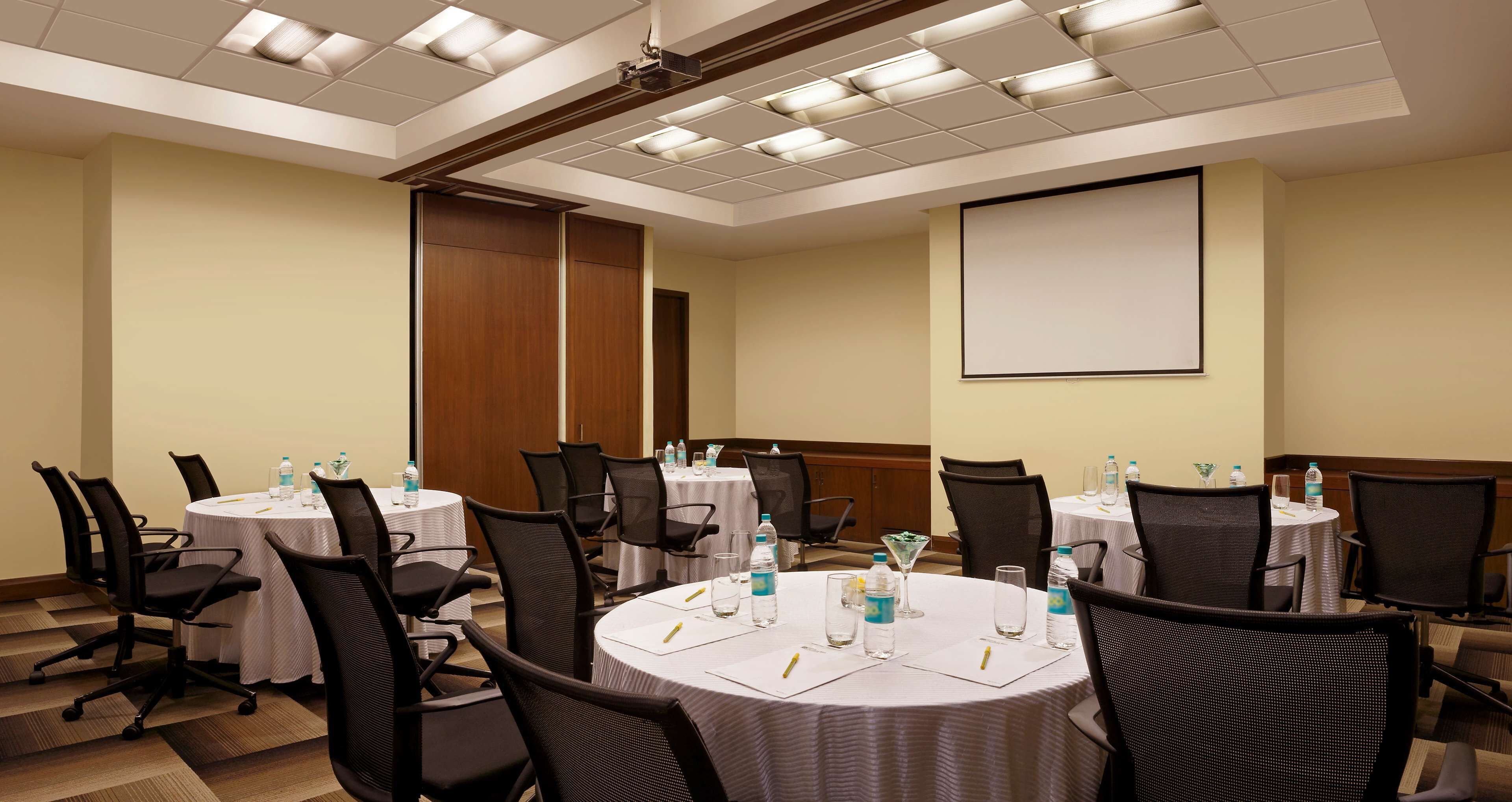 A conference room with multiple tables and chairs, equipped with water bottles and pens, and a projector screen on the wall.