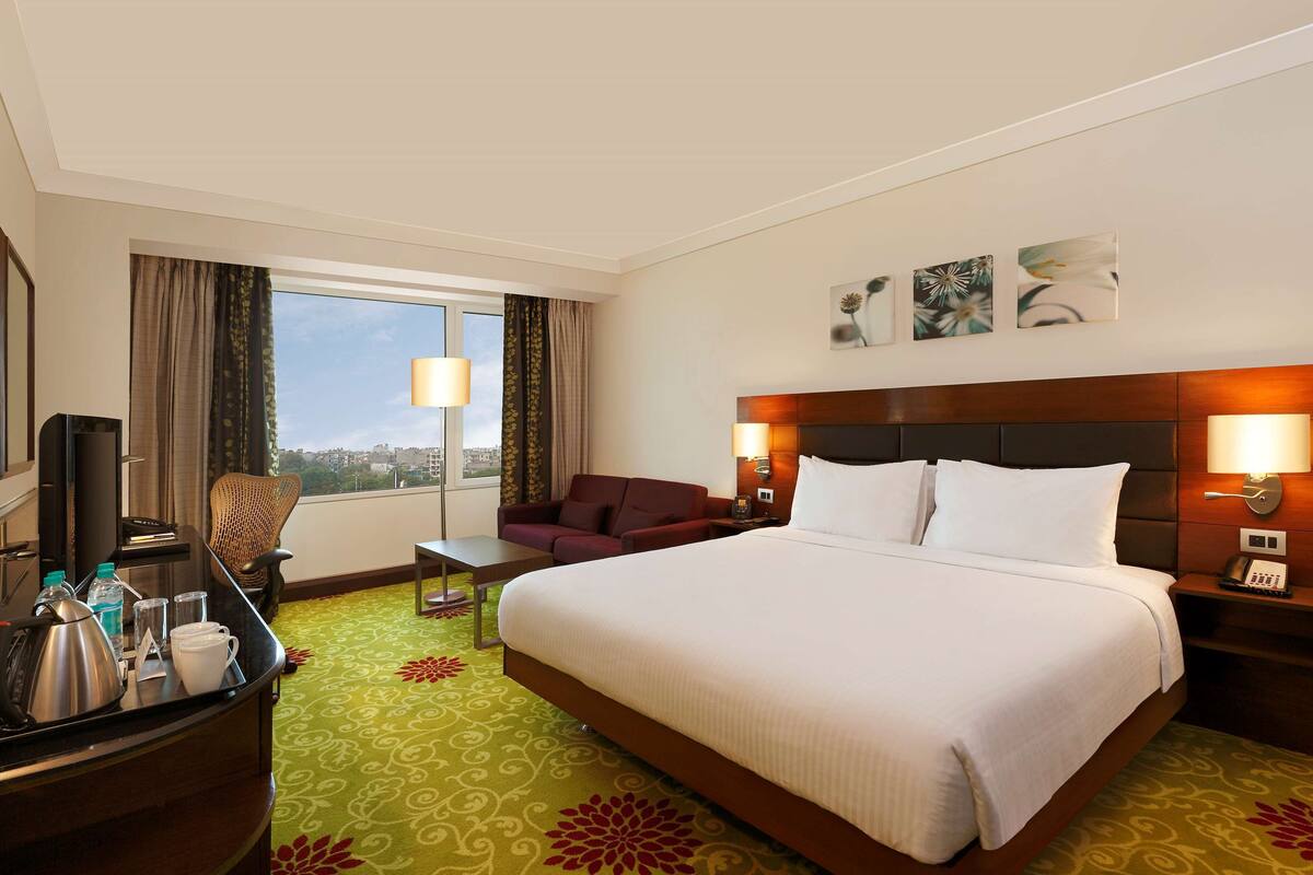 A hotel room with a large bed, a desk with a kettle, a chair, a TV, and a view of the city.