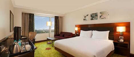 Ein Hotelzimmer mit einem großen Bett, einem Schreibtisch mit Wasserkocher, einem Sessel, einem Fernseher und Blick auf die Stadt.