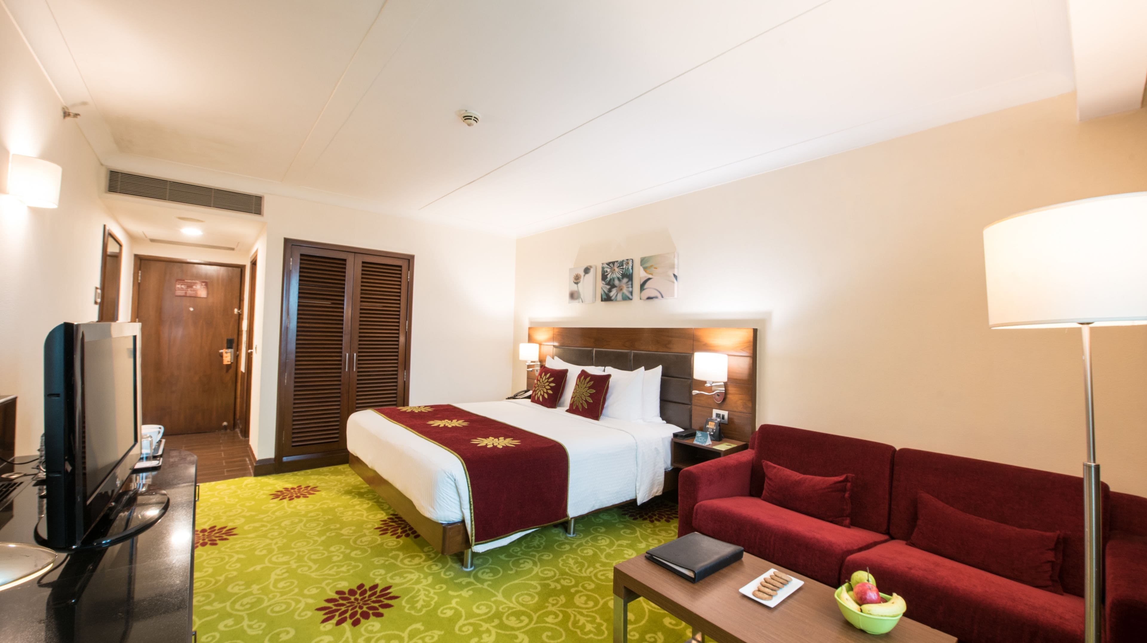A hotel room with a bed, a red sofa, a desk with a laptop, a TV, and a green carpet.